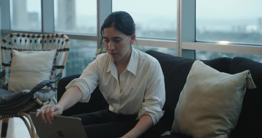 A businesswoman sits down on a couch in a modern office with a city view, opens her laptop, and stretches tiredly before continuing to work