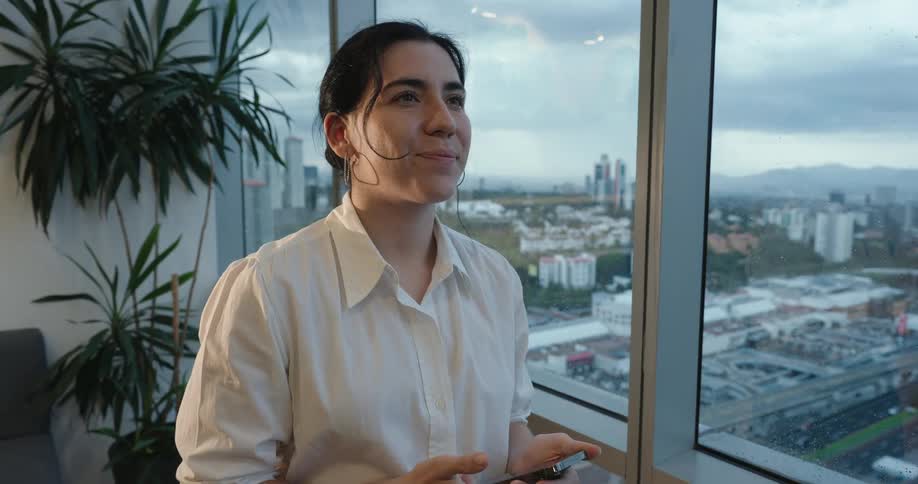 A young businesswoman smiles thoughtfully while looking out her office window at the cityscape after receiving good news on her smartphone