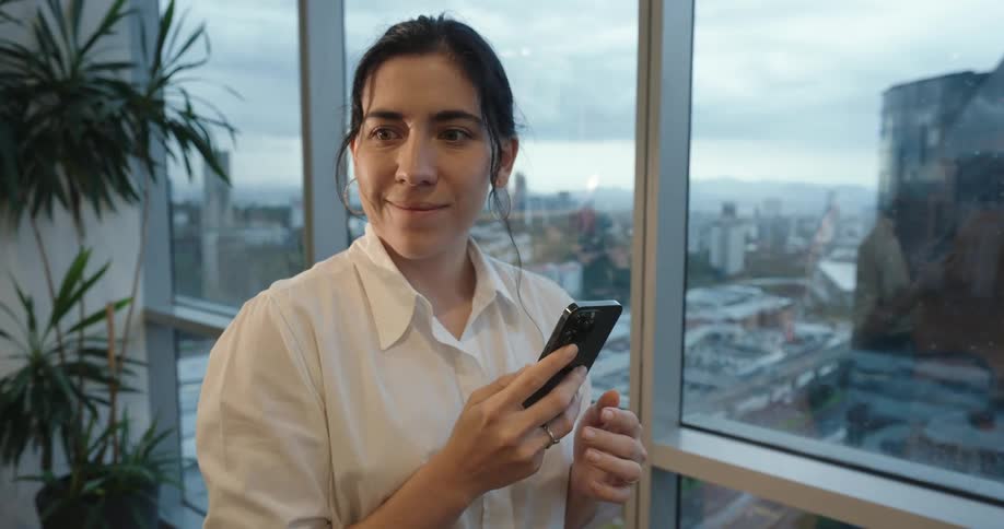 A young businesswoman's expression shifts from concern to a happy smile as she uses her smartphone in a modern high-rise office