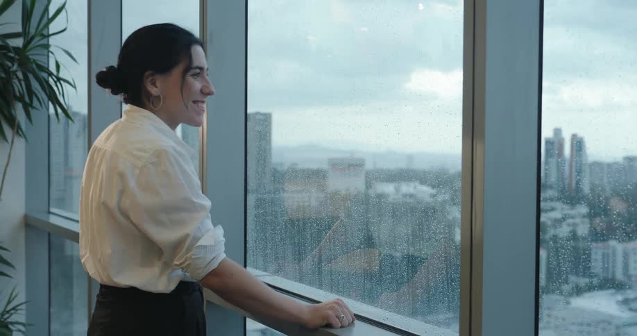 A happy businesswoman takes a break from work to enjoy the rainy day and the city view from her high-rise office window