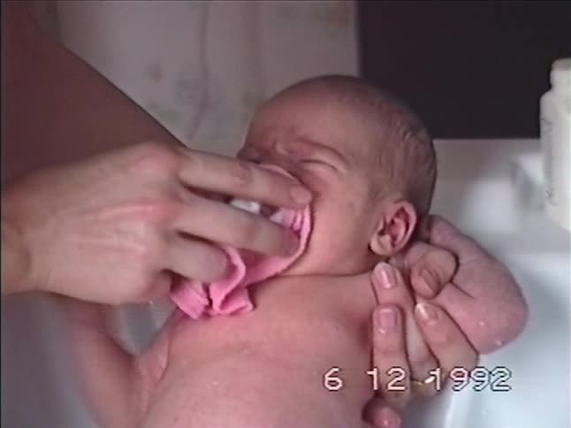 A parent's hands wash their newborn baby's face with a pink washcloth in a sink in this 1992 home video