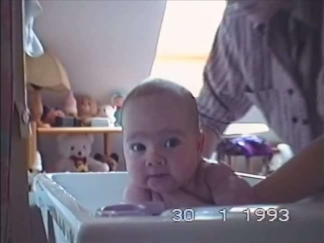 Close-up on a curious baby's face before zooming out to reveal them being gently bathed by a parent in this authentic VHS home movie from 1993