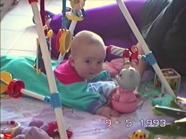 A cute baby in a colorful onesie plays on a playmat under a baby gym in this authentic home movie from 1993