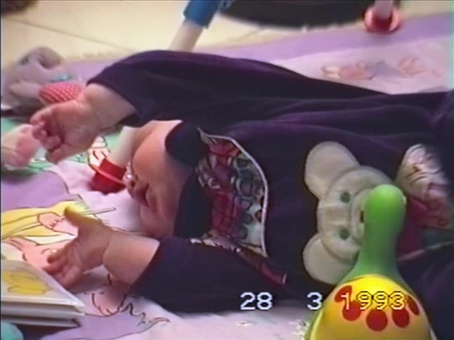 A cute baby lies on a colorful playmat, looking around curiously in this vintage home video from 1993
