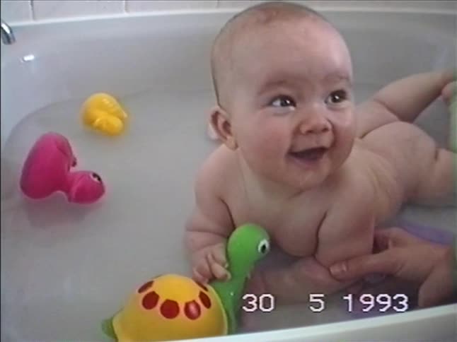 A cheerful baby lies on its stomach in the bathtub, smiling and laughing while playing with colorful toys in this vintage 1993 home video