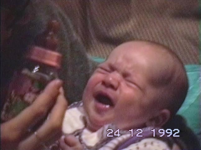 A woman calms her crying newborn baby by feeding them a bottle in a cozy living room on Christmas Eve 1992