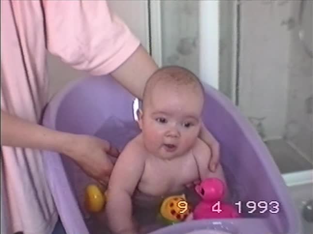 A mother tenderly washes her happy baby in a purple tub filled with colorful toys in an authentic 1993 home video