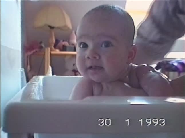 An adorable baby with big curious eyes gets a bath from their parent in a nostalgic home video from 1993
