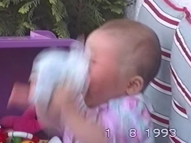 An adorable baby girl with chubby cheeks plays with her doll, shakes it, and then yawns in a retro 1993 home movie