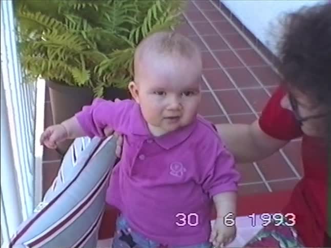 A vintage home video from 1993 shows a woman helping a cute baby stand on a patio cushion