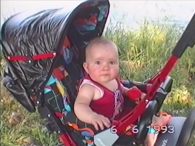 Authentic 1993 footage of an adorable baby in a red outfit sitting in a stroller by a lake, looking at the camera with wide eyes
