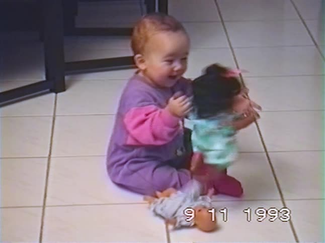 An adorable baby girl in a purple onesie sits on a tiled floor and happily plays with her dolls in a home video from 1993