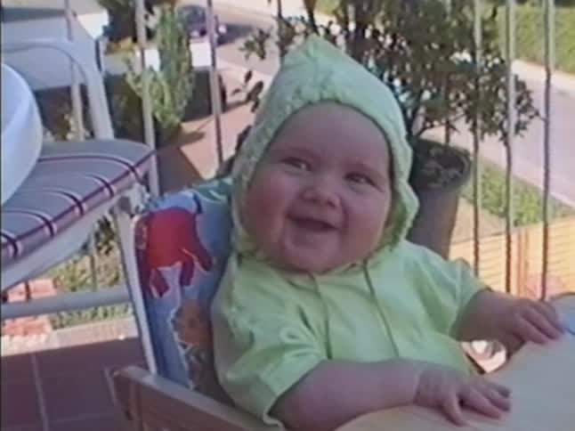 An adorable baby in a green pixie hat laughs uncontrollably while sitting in a high chair on a sunny balcony in a vintage home video