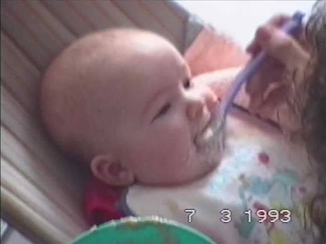 An over-the-shoulder shot captures a mother feeding her baby with a spoon in a bouncy chair, an authentic home video moment from 1993