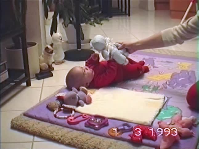 A vintage 1993 home video shows a baby in a red outfit lying on a play mat and interacting with a stuffed doll