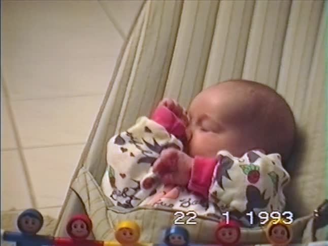 A cute newborn baby in a patterned onesie sits in a bouncer chair and explores its hands in a vintage home video from 1993