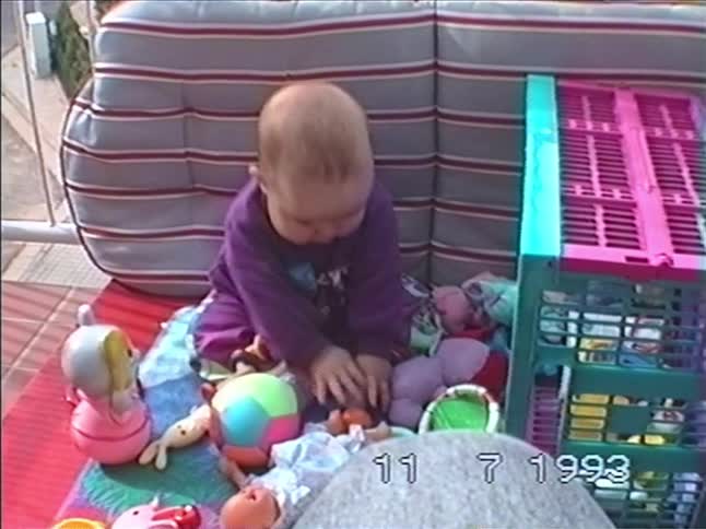 A vintage home video from 1993 shows a baby in a purple outfit playing with colorful toys on a patio