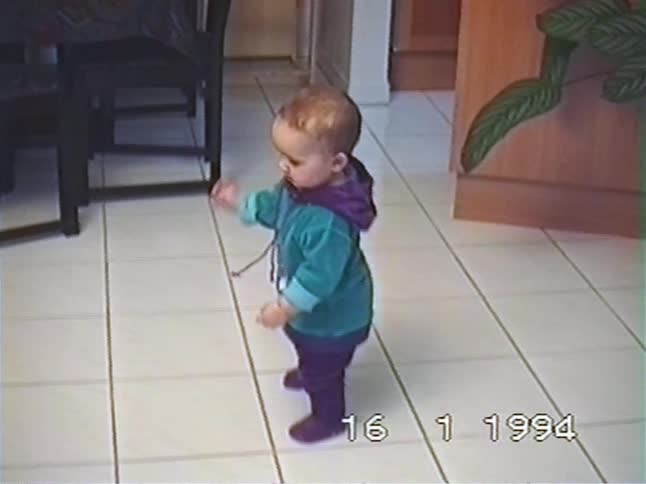 A cute baby in a colorful outfit dances and bounces on a tiled floor before sitting down in this authentic home movie from 1994