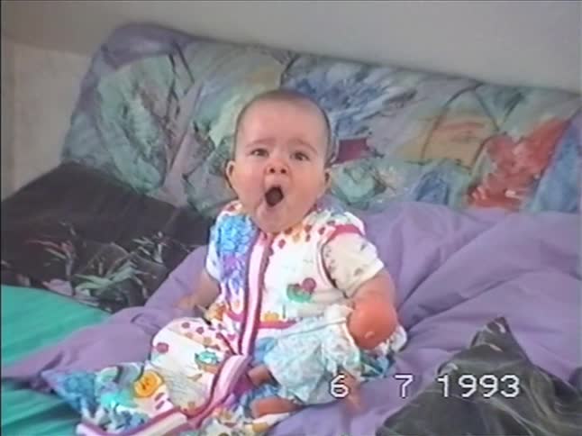 A sleepy baby in a colorful onesie sits on a bed and lets out a big yawn in this vintage home movie from 1993