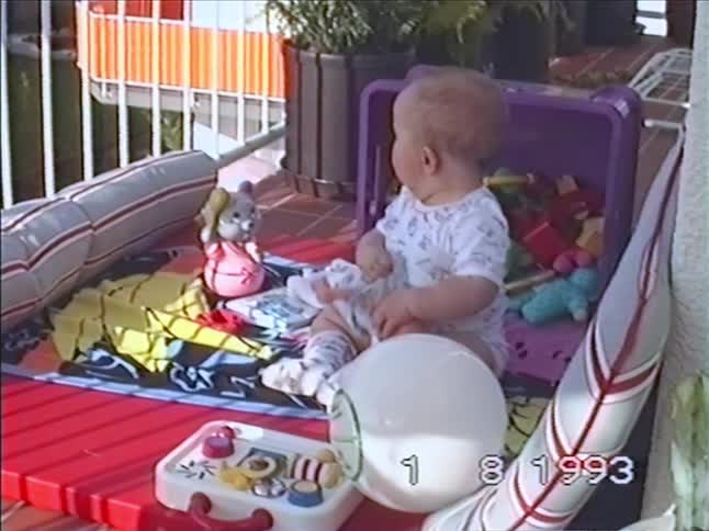 Vintage 1993 footage shows a baby playing with dolls and other toys in an outdoor playpen on a sunny day