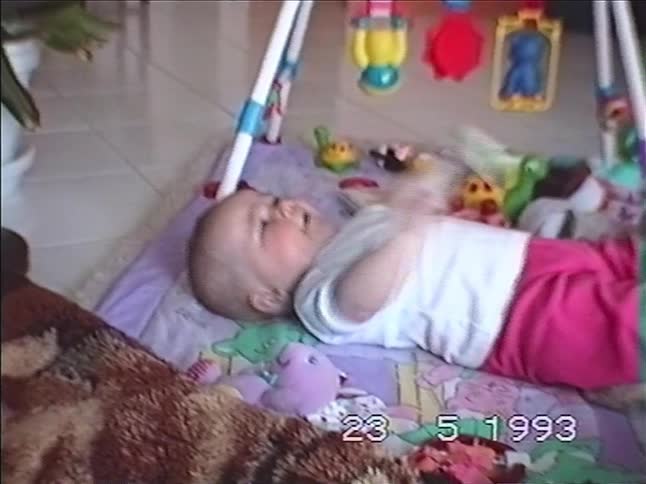 A happy baby lies on a playmat, kicking its legs and playing with toys in a vintage home video from 1993