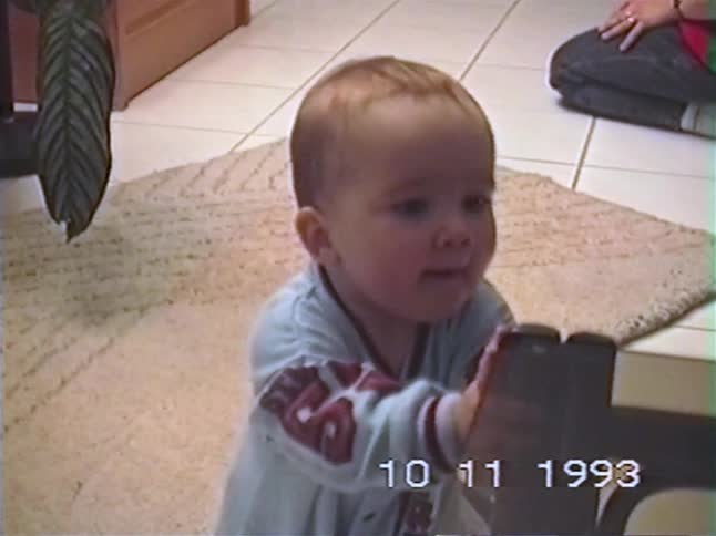 A cute baby learns to pull himself up on furniture and smiles at the camera in a home video from 1993