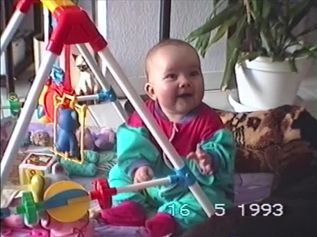Adorable baby in a colorful 90s outfit sits on the floor and bursts into a happy laugh while playing with a toy gym