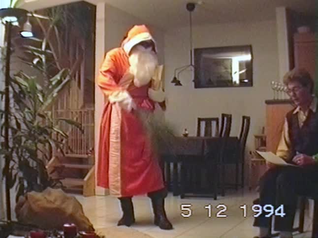 A person in a Saint Nicholas costume with a book and twigs entertains a woman during a holiday celebration in a 1994 home video