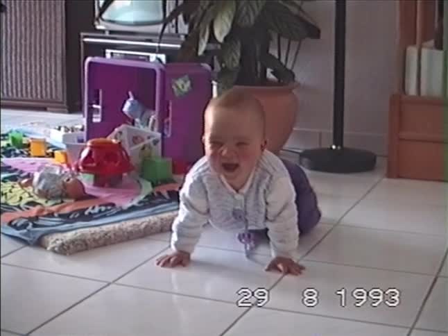 A cute baby on all fours on a tiled floor looks at the camera and lets out a big yawn before smiling in this home movie from 1993