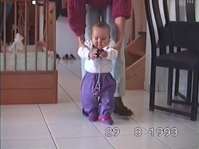 An adorable baby in purple pants takes wobbly first steps with the help of their parent in a heartwarming vintage 1993 home movie