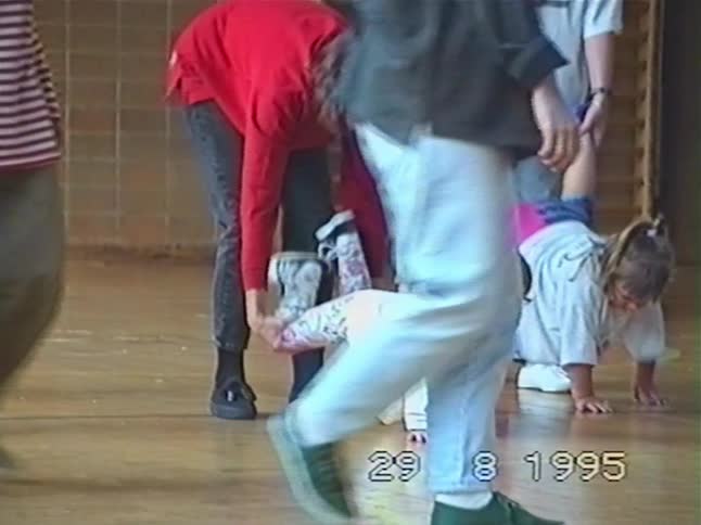 Vintage 1995 footage shows adults holding children's legs as they do a wheelbarrow walk race in a gymnasium