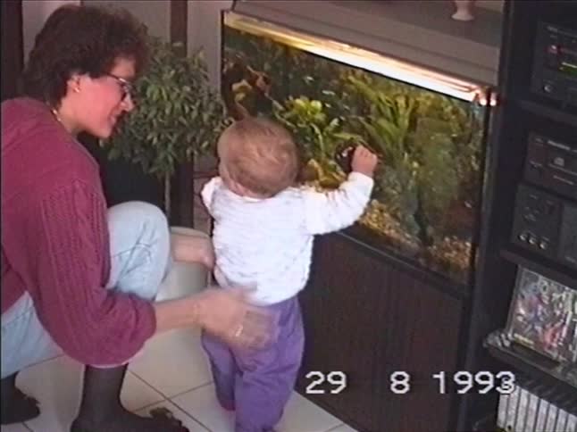 A mother helps her baby stand up to look into a large home aquarium in their living room, captured on VHS in 1993