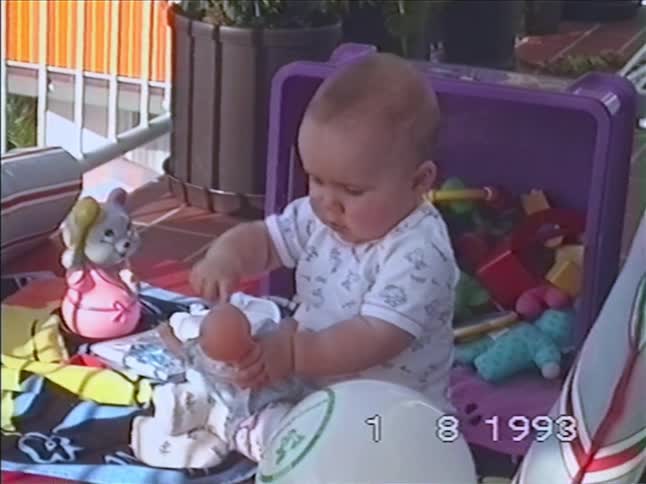 A cute baby sits on a blanket outdoors surrounded by toys and plays with a doll in this vintage home video from 1993