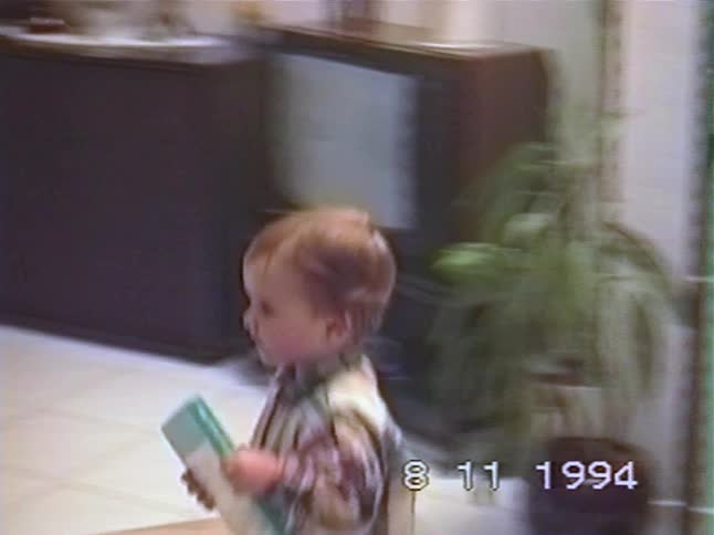 An adorable toddler proudly holds a book and walks through the living room in a 1994 home video