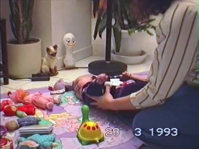 A mother playfully helps her baby sit up and then lays them down on a playmat in this archival home video from 1993