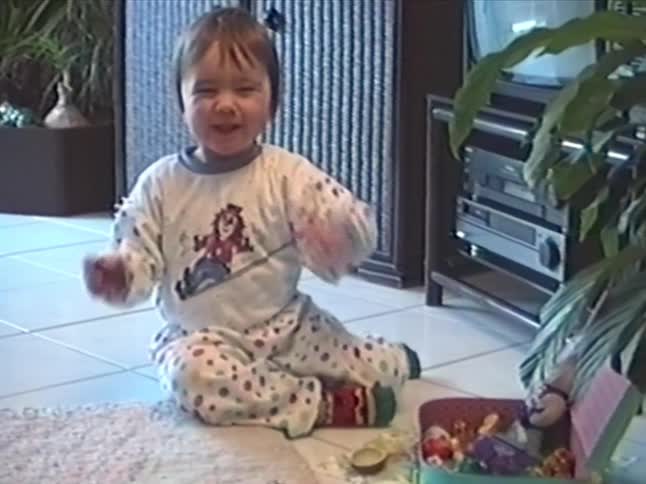 An adorable toddler in polka-dot pajamas happily eats a snack and waves at the camera while sitting on the floor