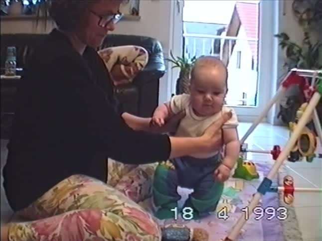 A mother lovingly supports her baby as they practice standing and bouncing on a colorful playmat in a 1993 home video