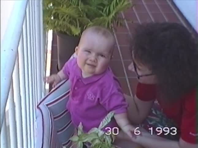 Adorable vintage 1993 footage shows a baby sitting with their mother on a tiled patio, curiously playing with a potted plant