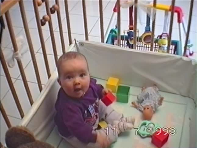 An adorable baby in a purple shirt sits in a playpen, playing with blocks before looking up at the camera with wide eyes and an open mouth in this 1993 home video