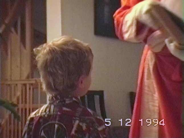 A young boy stands before Saint Nicholas, who reads from his large book, in a charming home video from 1994