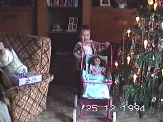 A sweet toddler enjoys their new doll and stroller next to the Christmas tree in a nostalgic home video from 1994