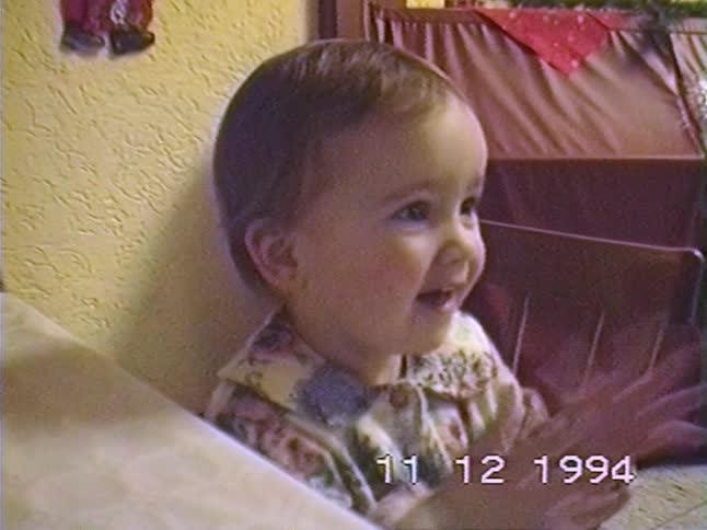 An adorable baby from a 1994 home video happily claps and smiles while sitting at a table