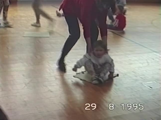 A mother stands behind her toddler who is sitting on a mat on a gym floor during a group activity in this vintage 1995 footage