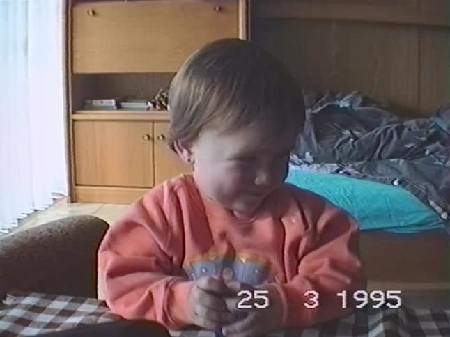 A toddler sits at a checkered table and plays intently with a toy in this authentic home video from 1995