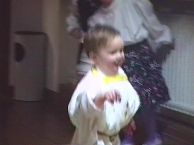 A cute toddler wearing a white costume with a yellow sash smiles and walks around at a family gathering