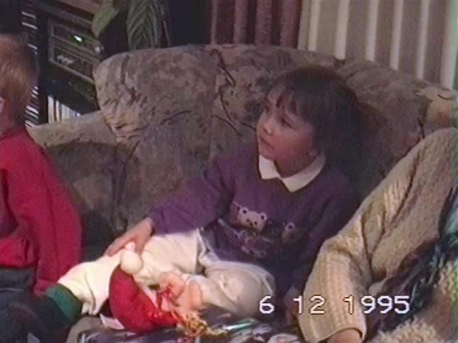 A little girl in a purple sweater sits on a couch looking up with curiosity in a home video from 1995