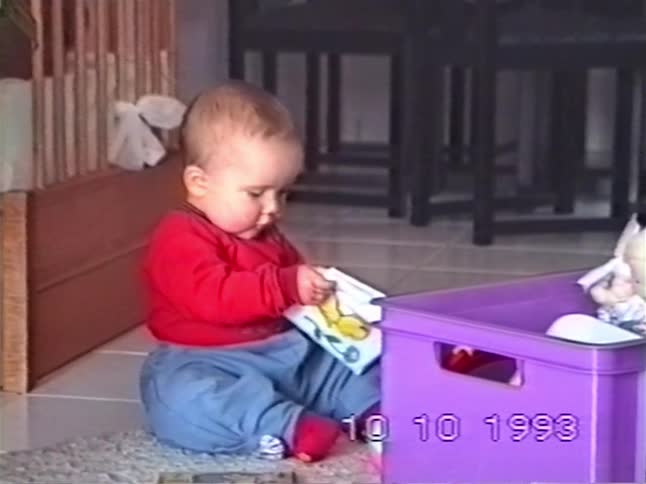 An adorable baby in a red shirt sits on the floor and curiously looks through a picture book from their toy box in this nostalgic 1993 home video