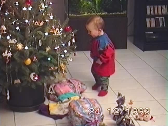 A cute toddler is mesmerized by a decorated Christmas tree with presents on Christmas Eve in 1993
