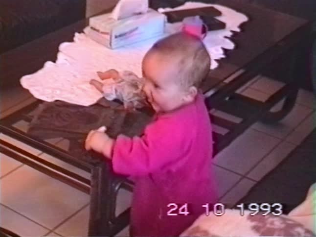 An adorable baby girl in a pink outfit stands by a coffee table, playing with a doll and smiling brightly at the camera in a nostalgic 1993 home video