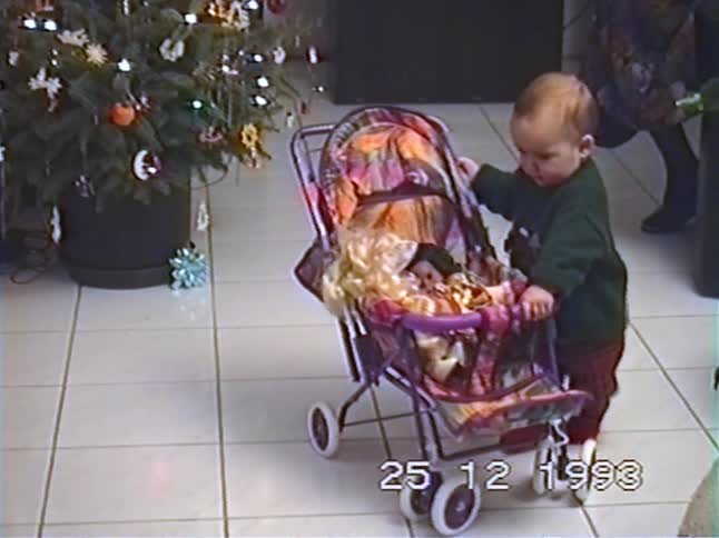 Vintage home movie captures a cute toddler playing with a new doll stroller next to the Christmas tree in 1993